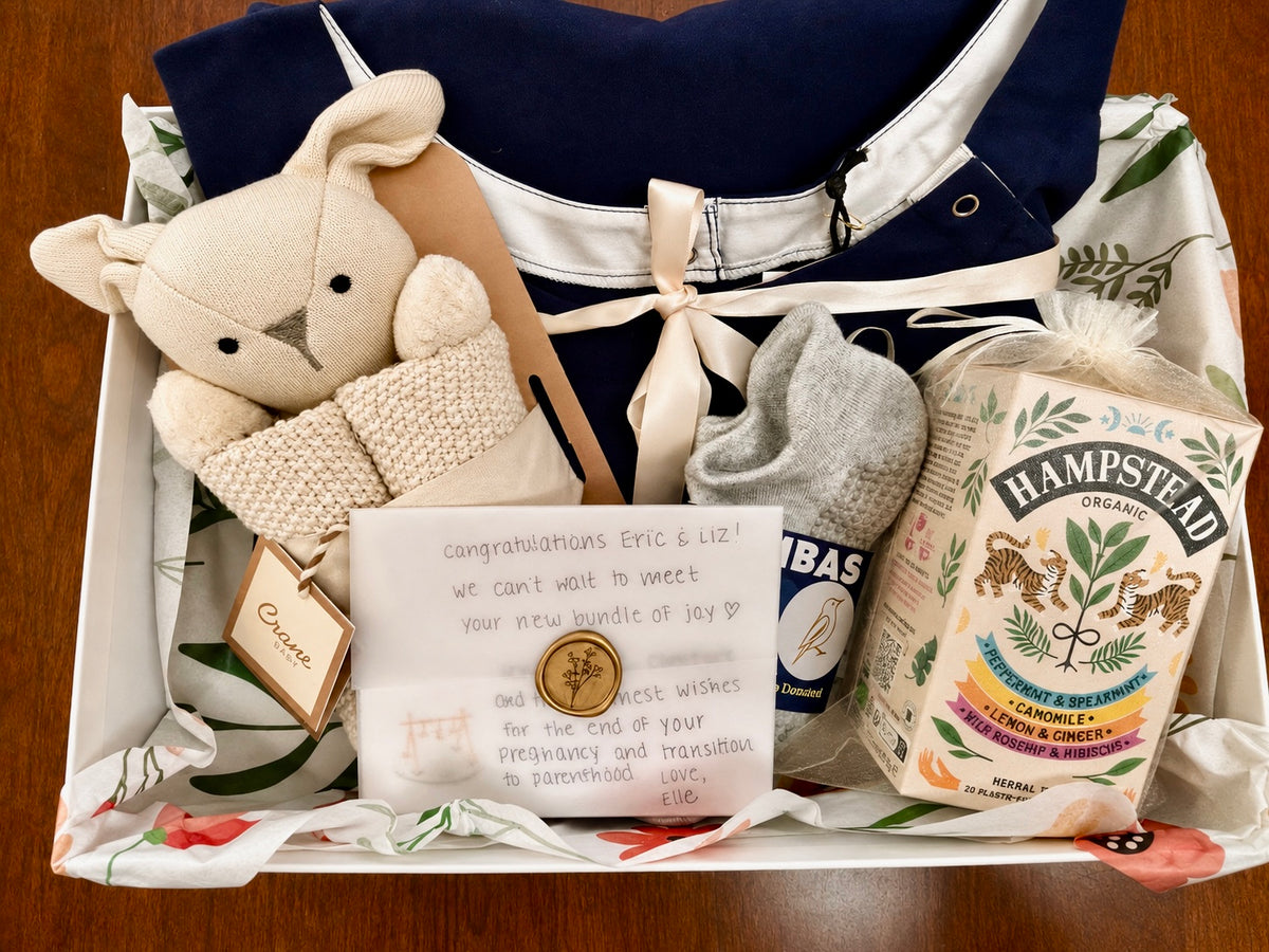 A maternity and baby gift box filled with curated items: organic cotton maternity gown, Hampstead Organic tea, Bombas gripper socks, Organic Baby Lovey/Blanket and a handwritten congratulations card.