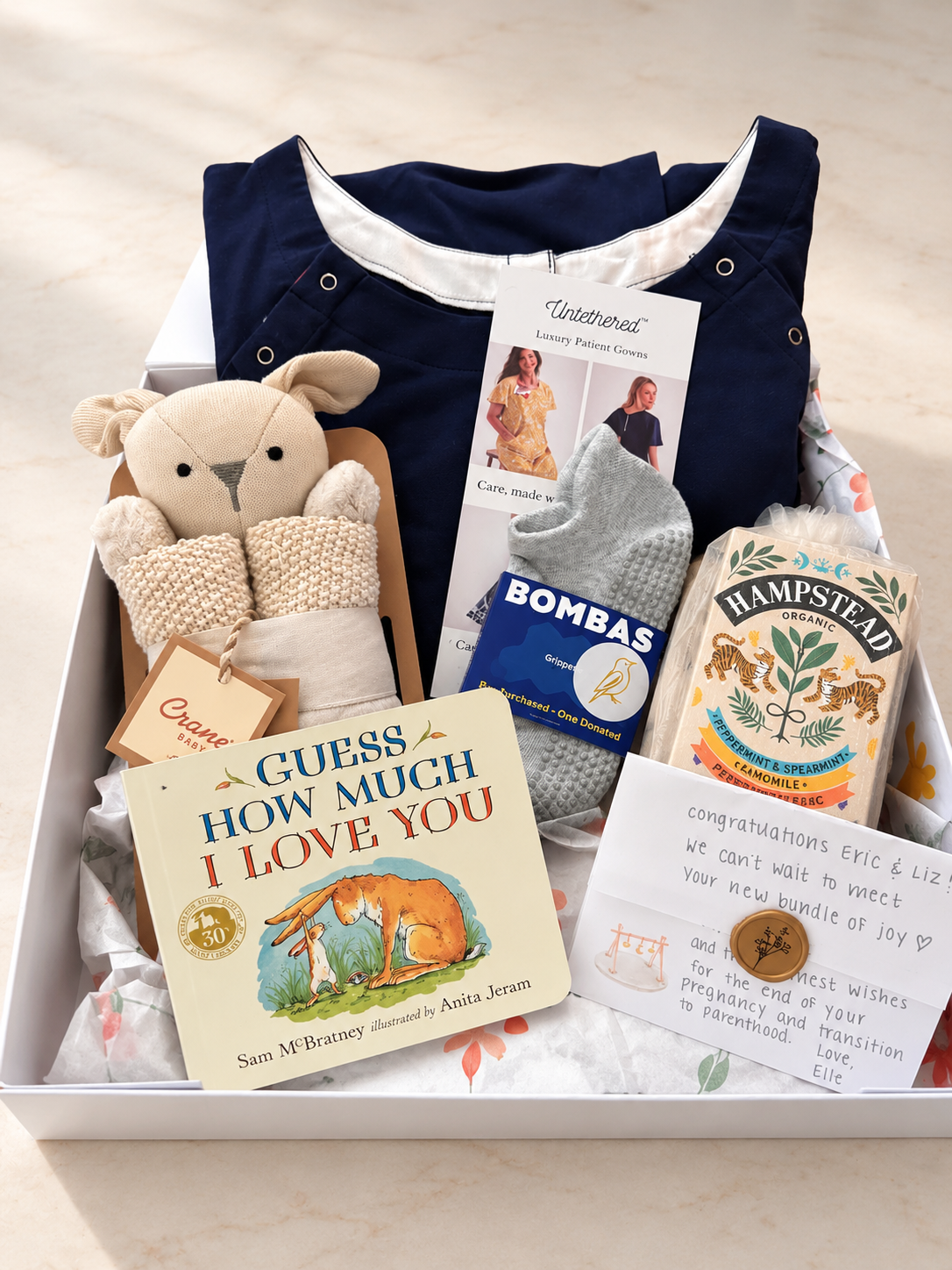 A baby gift box filled with curated items including a navy organic cotton labor gown, a plush organic lovey blanket, a book titled 'Guess How Much I Love You', Hampstead organic tea, Bombas gripper socks and a congratulations card, arranged in a white box on a light surface.